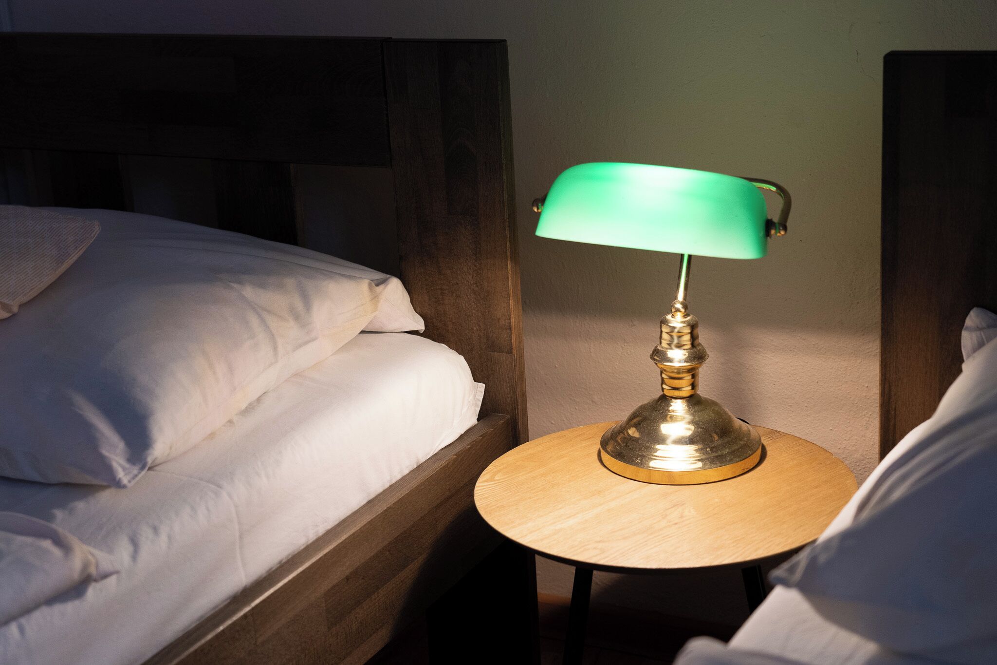 A single bed with white bedding, a round wooden nightstand with a green lamp, and a wooden headboard.