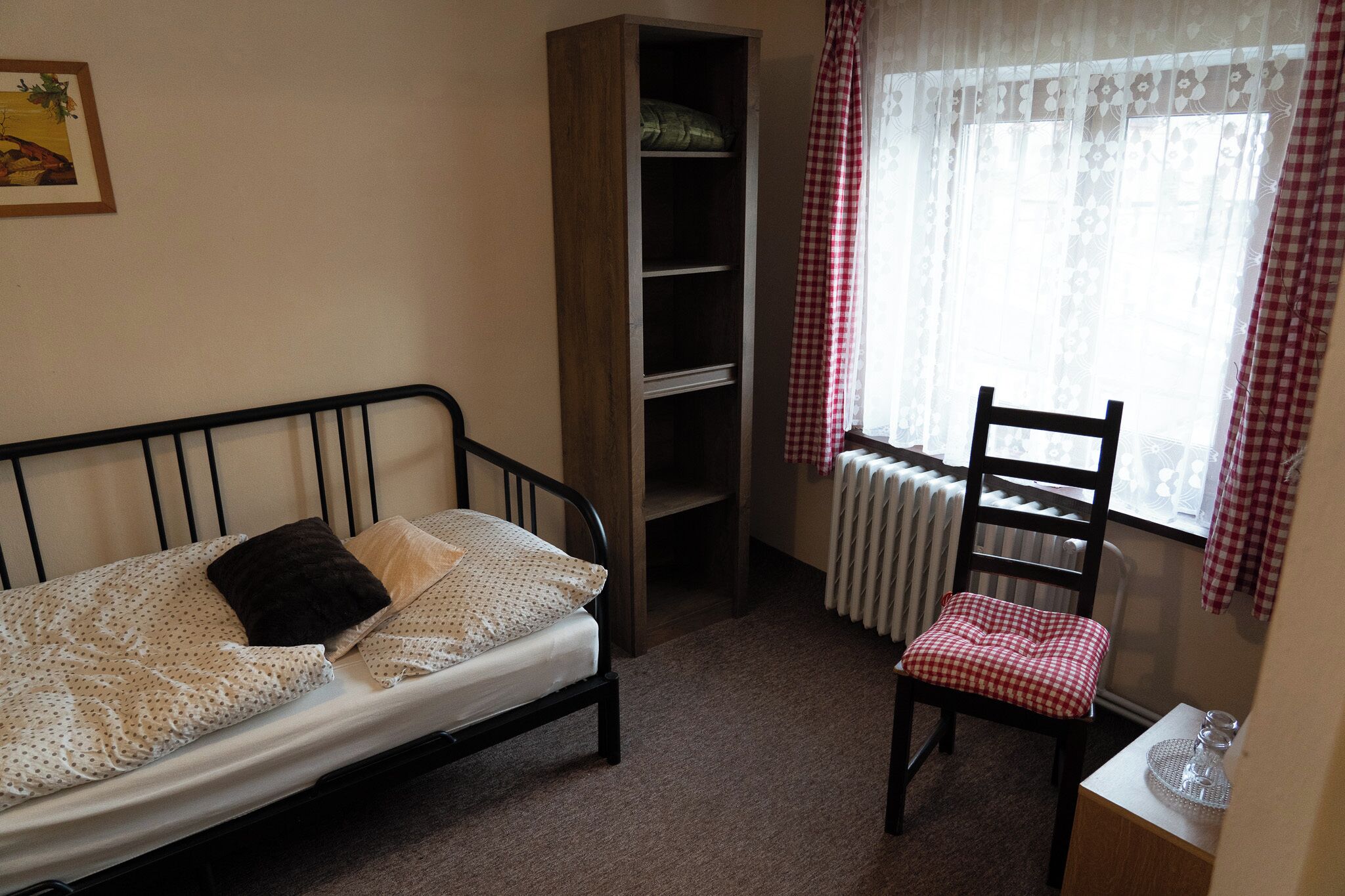 A small, neatly arranged bedroom with a single bed, a chair, a bookshelf, and a radiator near a window with checkered curtains.