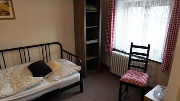 A small, neatly arranged bedroom with a single bed, a chair, a bookshelf, and a radiator near a window with checkered curtains.