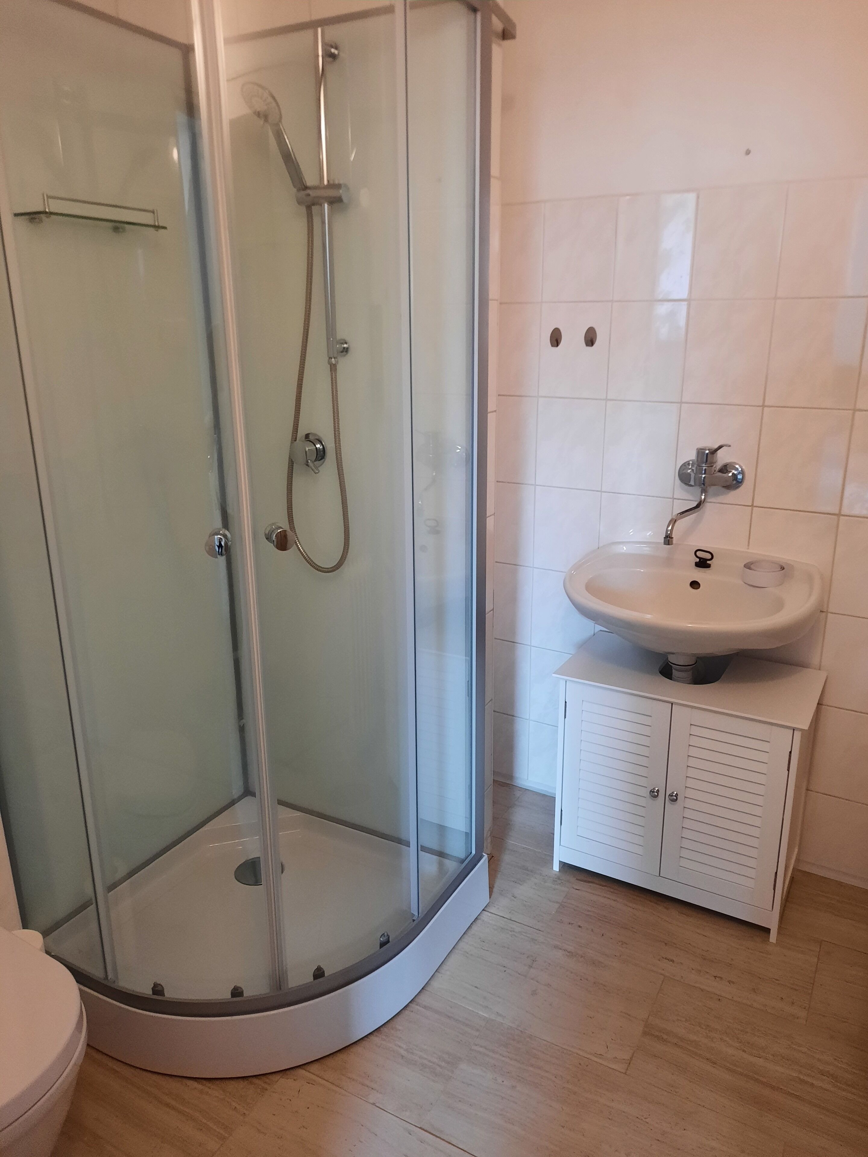 Una ducha con cerramiento de vidrio, con un cabezal de ducha manual y un grifo de pared. Junto a la ducha, un lavabo blanco de una sola cubeta con un mueble debajo.
