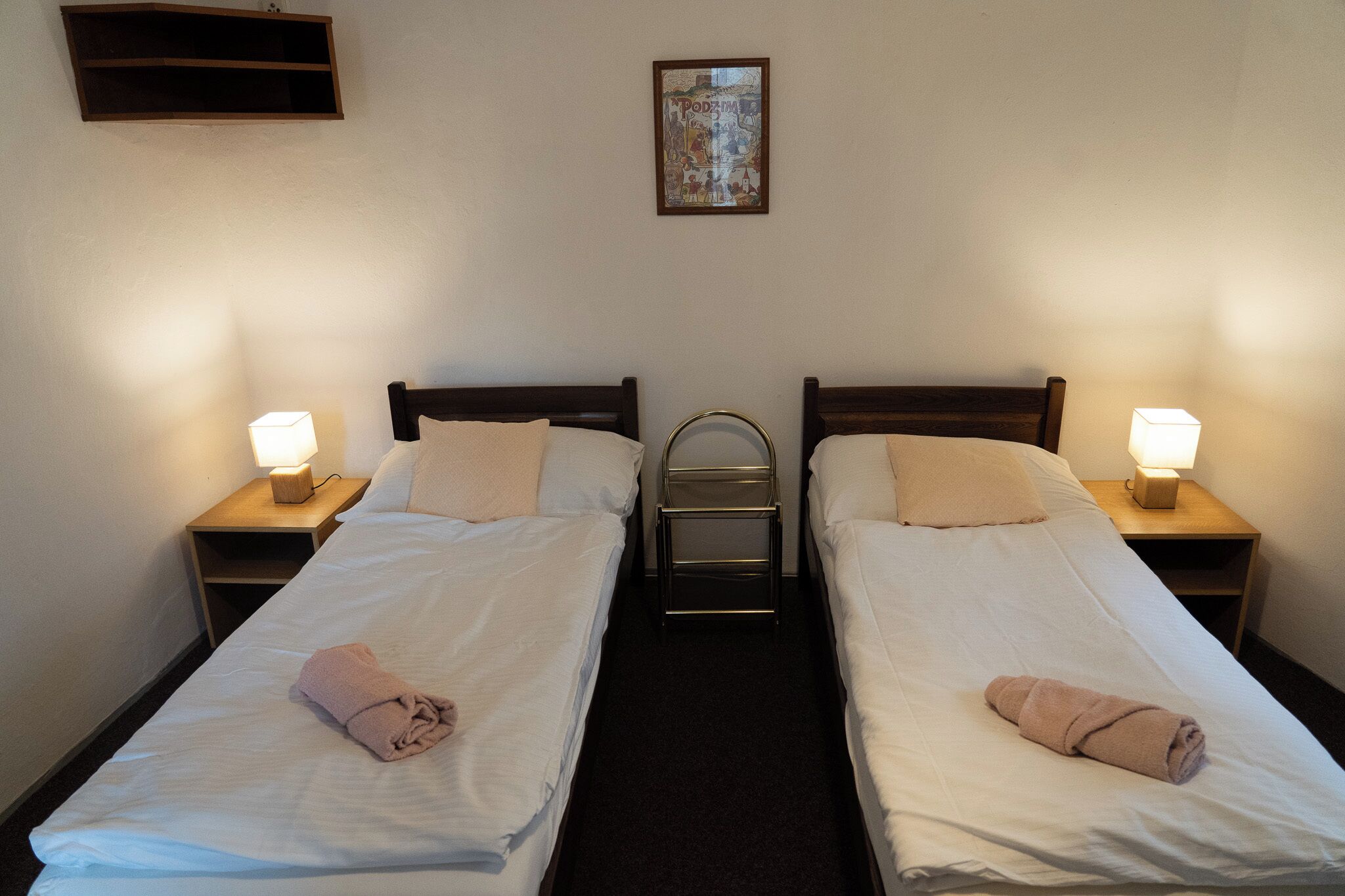 Ein Hotelzimmer mit zwei Einzelbetten, jeweils mit Nachttisch und Lampe. Über dem linken Bett befindet sich eine kleine Ablagefläche und an der Wand ein gerahmtes Bild.