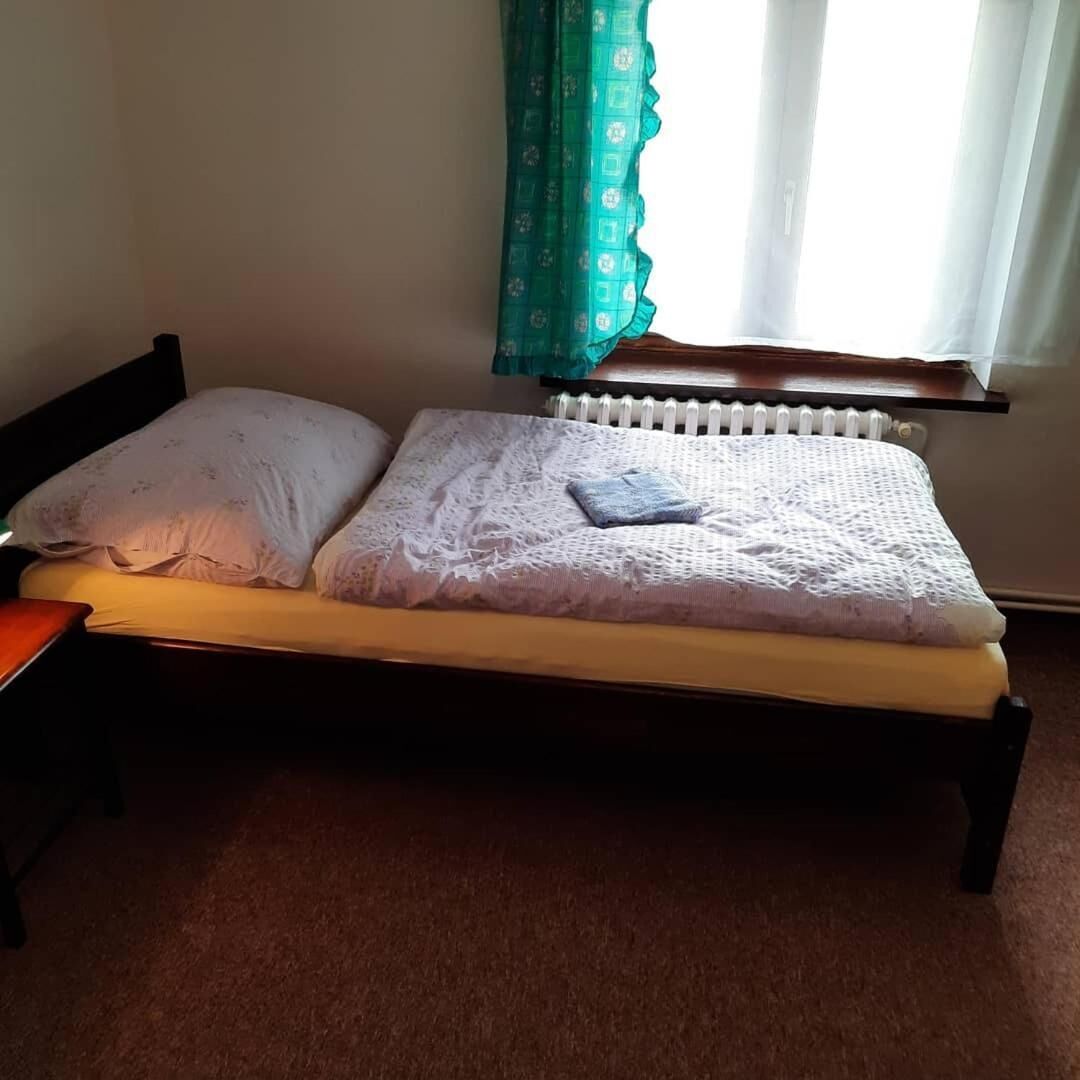 Una cama individual con colchón blanco y almohada azul, cabecera de madera, mesita de noche y ventana con cortina.