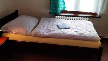 Una cama individual con colchón blanco y almohada azul, cabecera de madera, mesita de noche y ventana con cortina.