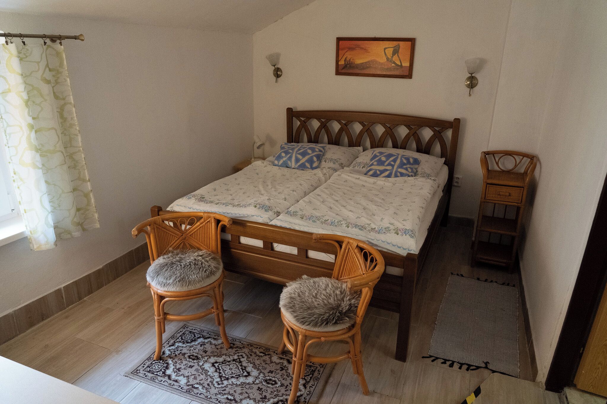 Ein Schlafzimmer mit einem hölzernen Bett, zwei Stühlen, einem kleinen Holztisch, einem Teppich und einem Gemälde an der Wand.