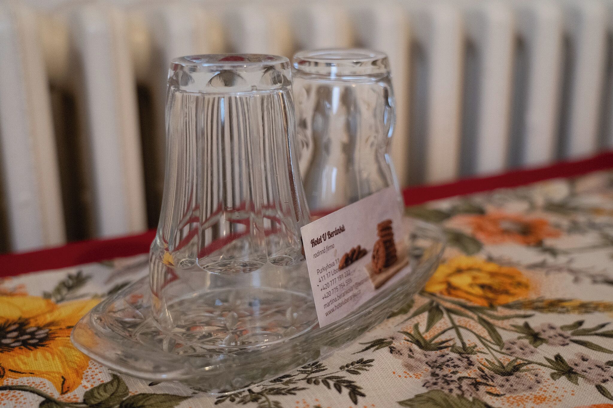 A clear glass decanter and a glass pitcher on a floral tablecloth with a label indicating 'Hotel U Berlins'.
