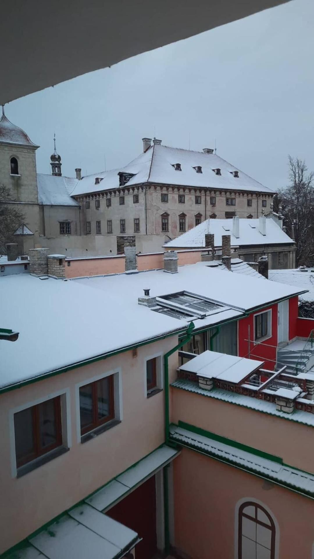 A building with a snow-covered roof, a red building in the foreground, and a church with a dome in the background.