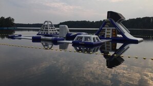 An outdoor water park with inflatable play structures, including a slide and climbing wall, set against a calm lake with trees in the background.