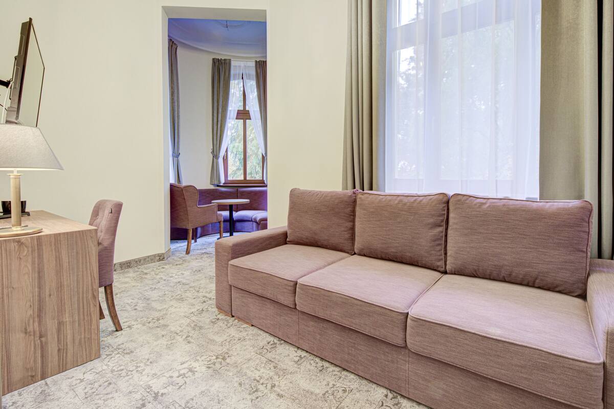 Ein Wohnzimmer mit einem beigefarbenen Sofa, einem Holztisch und einer Lampe. Ein Fenster mit Vorhängen bietet Blick ins Grüne.