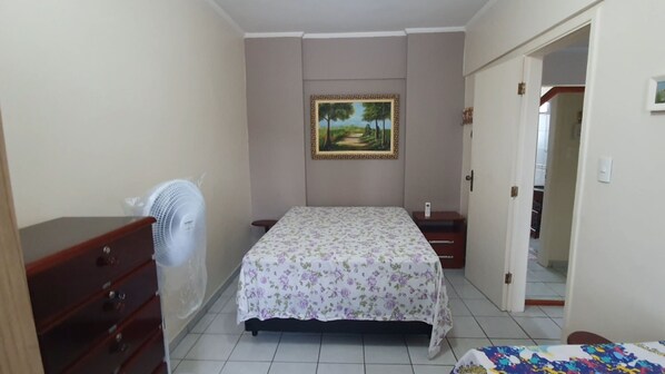 A bedroom with a bed, a dresser, a nightstand, a wall-mounted fan, and a door leading to another room.