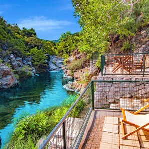 A clear view of a river with rocky banks, surrounded by lush greenery. There is a wooden deck with a chair and a fence. - Yiva Kazdaglari (Edremit)