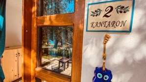 A wooden door with a window revealing a view of a garden and a wooden bench. A sign on the wall reads "2 KANTIARON" with a decorative blue owl ornament. - Yiva Kazdaglari (Edremit)