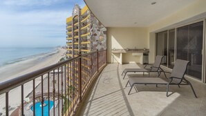 A balcony with a view of the beach, a pool, and a dining area with chairs.