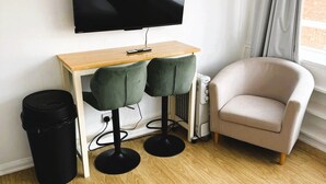 Un salón moderno con un escritorio de madera, dos taburetes verdes, un sillón color beige, un cubo de basura negro y un televisor de pantalla plana montado en la pared.