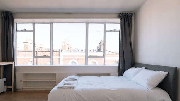 Una habitación de hotel con un ventanal grande, una cama con ropa de cama blanca, una mesita de noche y un radiador.