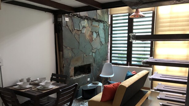 Ein gemütliches Wohnzimmer mit Kamin aus Stein, einem Esstisch mit weißen Schalen, einer Couch mit Kissen und einem Fenster mit Jalousien.