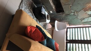 Ein Wohnzimmer mit Steindecke, einer Couch mit Kissen, einem kleinen Tisch und einem Fenster mit Jalousien.
