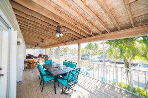 Un espacio al aire libre techado con techo de madera, una mesa con sillas y vistas a palmeras y un estacionamiento.