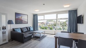 A modern living room with a sofa, a dining table, and chairs. There is a large window with curtains, a flat-screen TV mounted on the wall, and a painting on the wall. - Blue Shell With Private Parking Garage (Zandvoort)