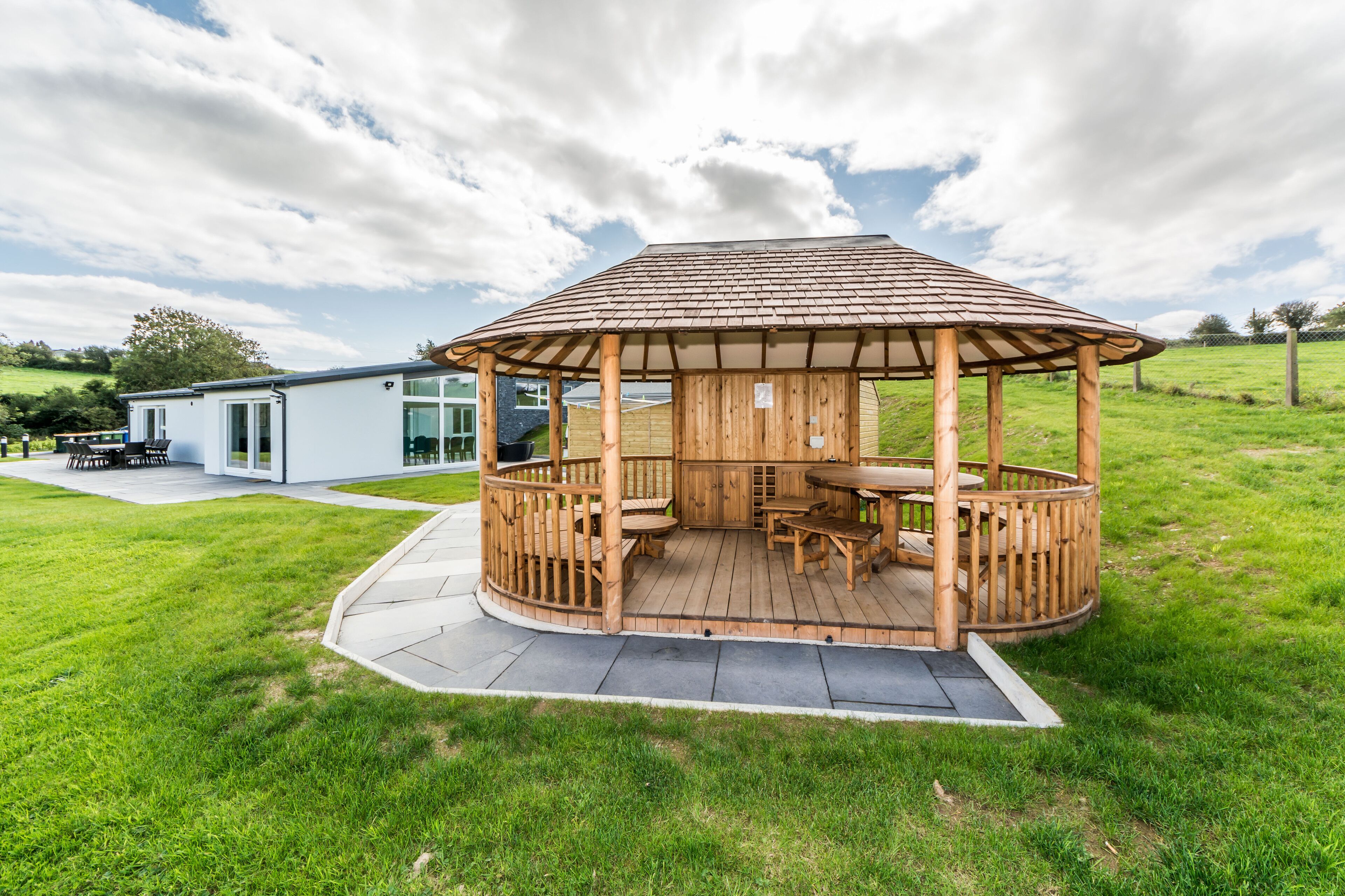A wooden gazebo with a tiled roof, surrounded by a grassy lawn and a modern building in the background.
