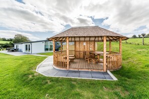 En træpavillon med et tag af fliser, omgivet af en græsplæne og en moderne bygning i baggrunden.