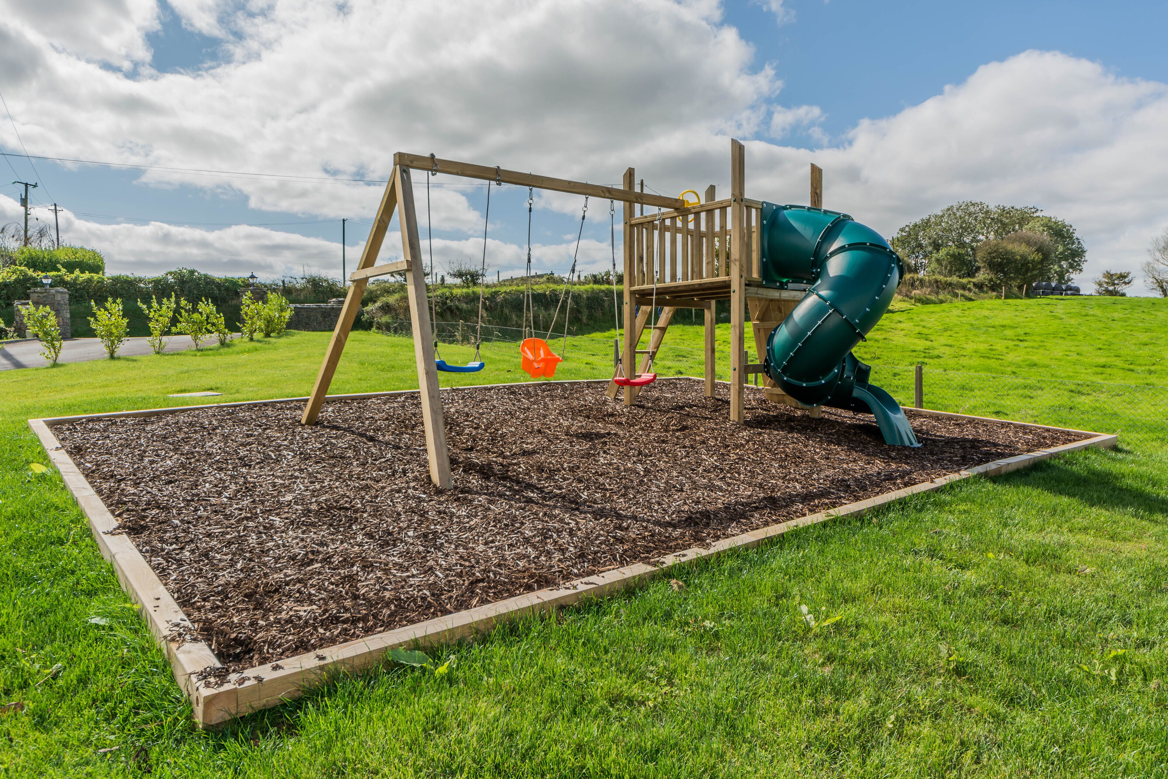 Ein hölzerner Spielplatz mit Schaukel und Rutsche, umgeben von einer Rasenfläche und einem Blumenbeet.