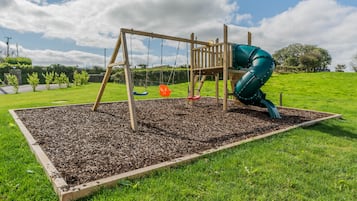 Ein hölzerner Spielplatz mit Schaukel und Rutsche, umgeben von einer Rasenfläche und einem Blumenbeet.