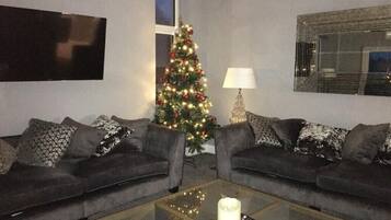 A living room with two grey sofas, a glass coffee table with a remote control, a Christmas tree, and a lamp.