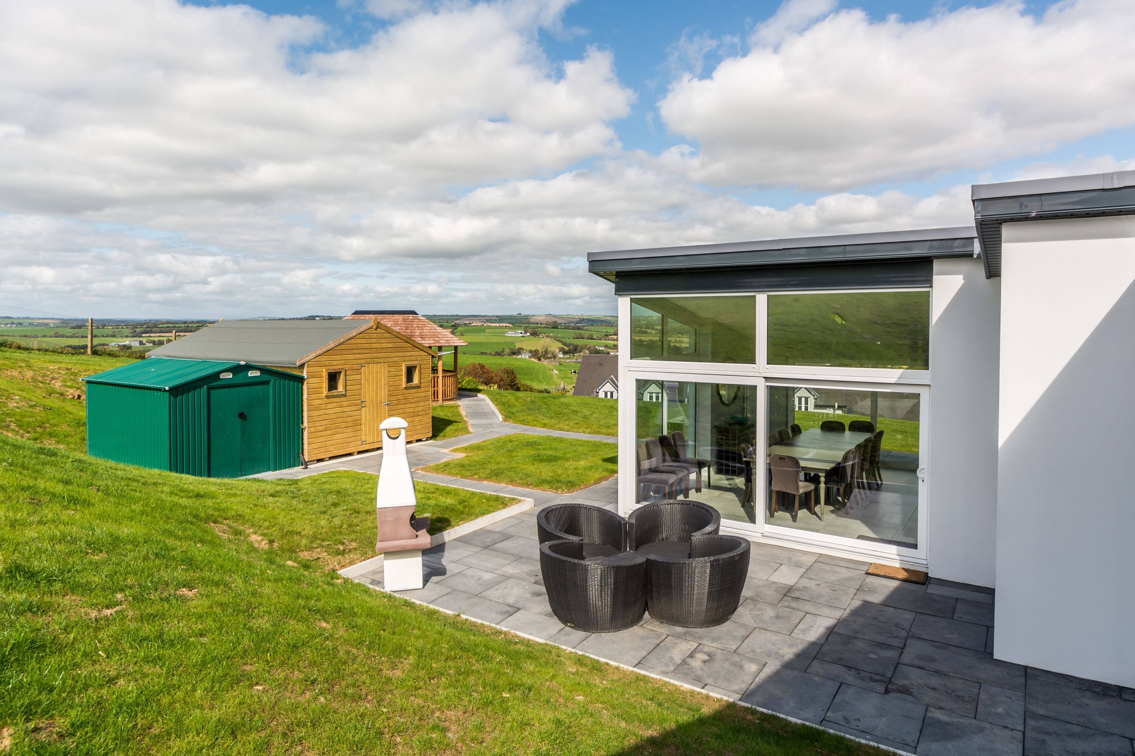 A modern house with a patio area, a green shed, and a wooden cabin in the background.