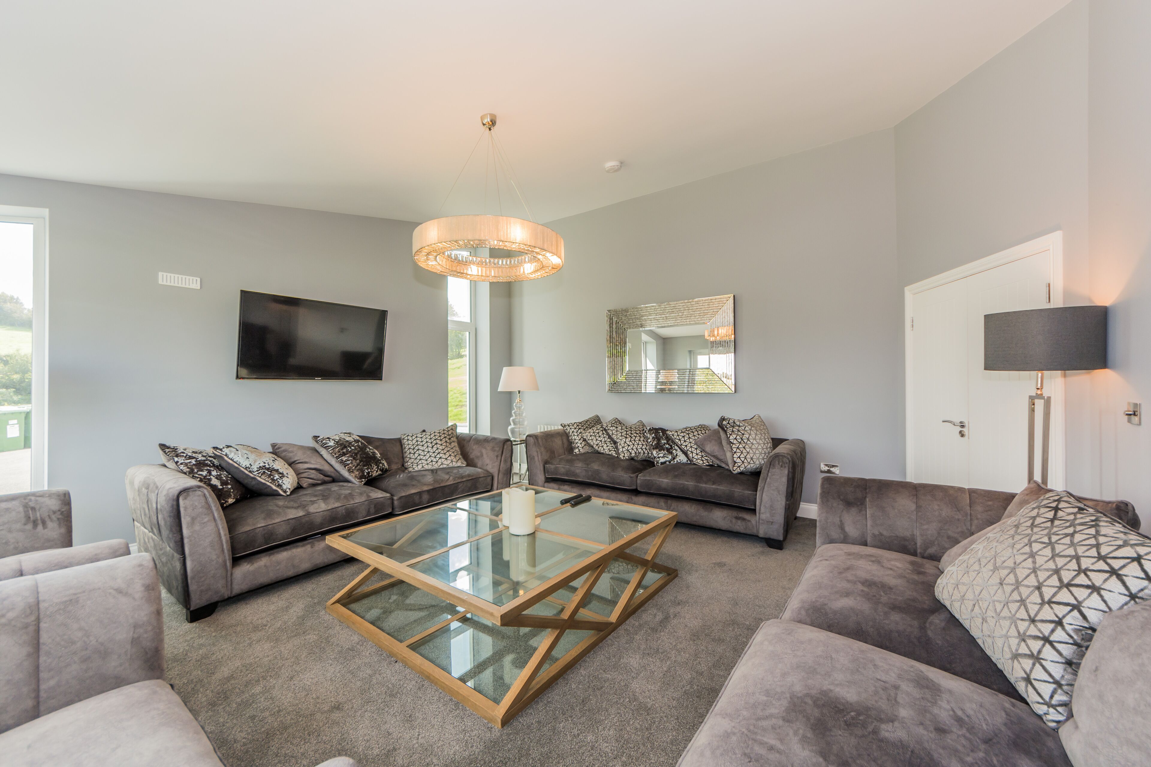 A modern living room with grey sofas, a glass coffee table, a mounted TV, and a large mirror.
