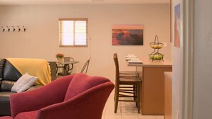 Un salon avec un fauteuil rouge, un coin repas avec une table et des chaises, et une cuisine avec un panier de fruits.