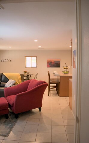 A living room with a red armchair, a dining area with a table and chairs, and a kitchen area with a fruit basket.