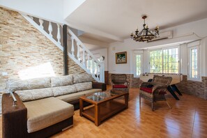 A spacious living room with a wooden floor, a sofa, a coffee table, and a wicker chair. A staircase with white balusters leads to another level.