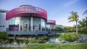 Un bâtiment moderne à la structure circulaire, doté d’un balcon avec des rampes violettes et agrémenté d’un élément décoratif aquatique en premier plan.