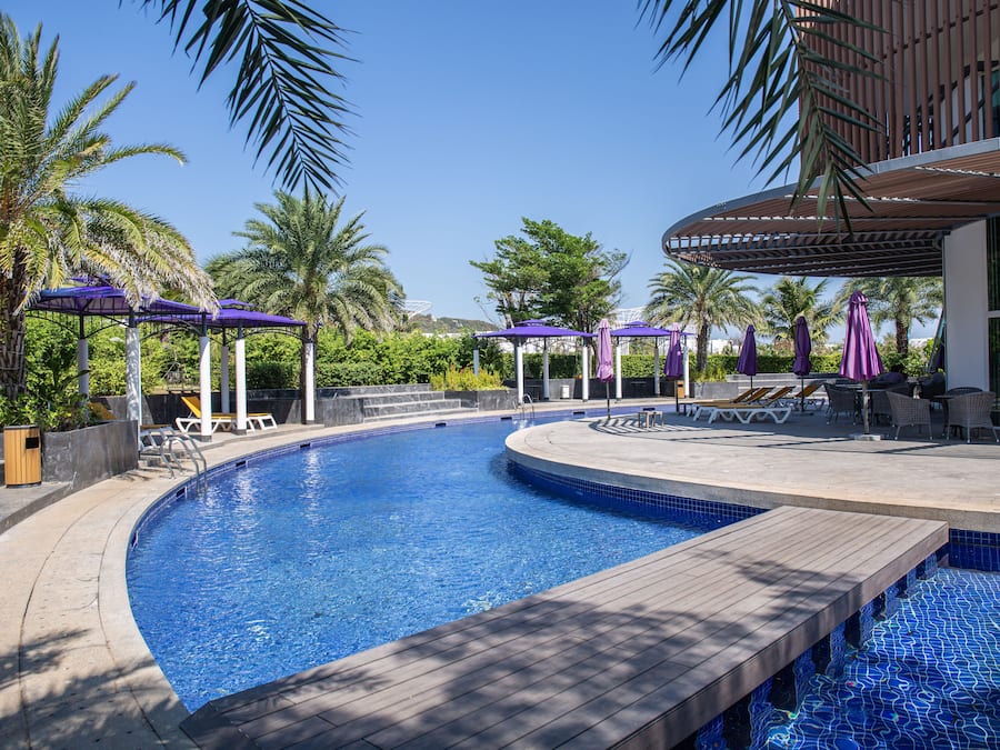 Una piscina con sillones de descanso y sombrillas, rodeada de palmeras y una zona de estar cubierta.