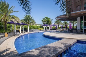 Una piscina con sillones de descanso y sombrillas, rodeada de palmeras y una zona de estar cubierta.