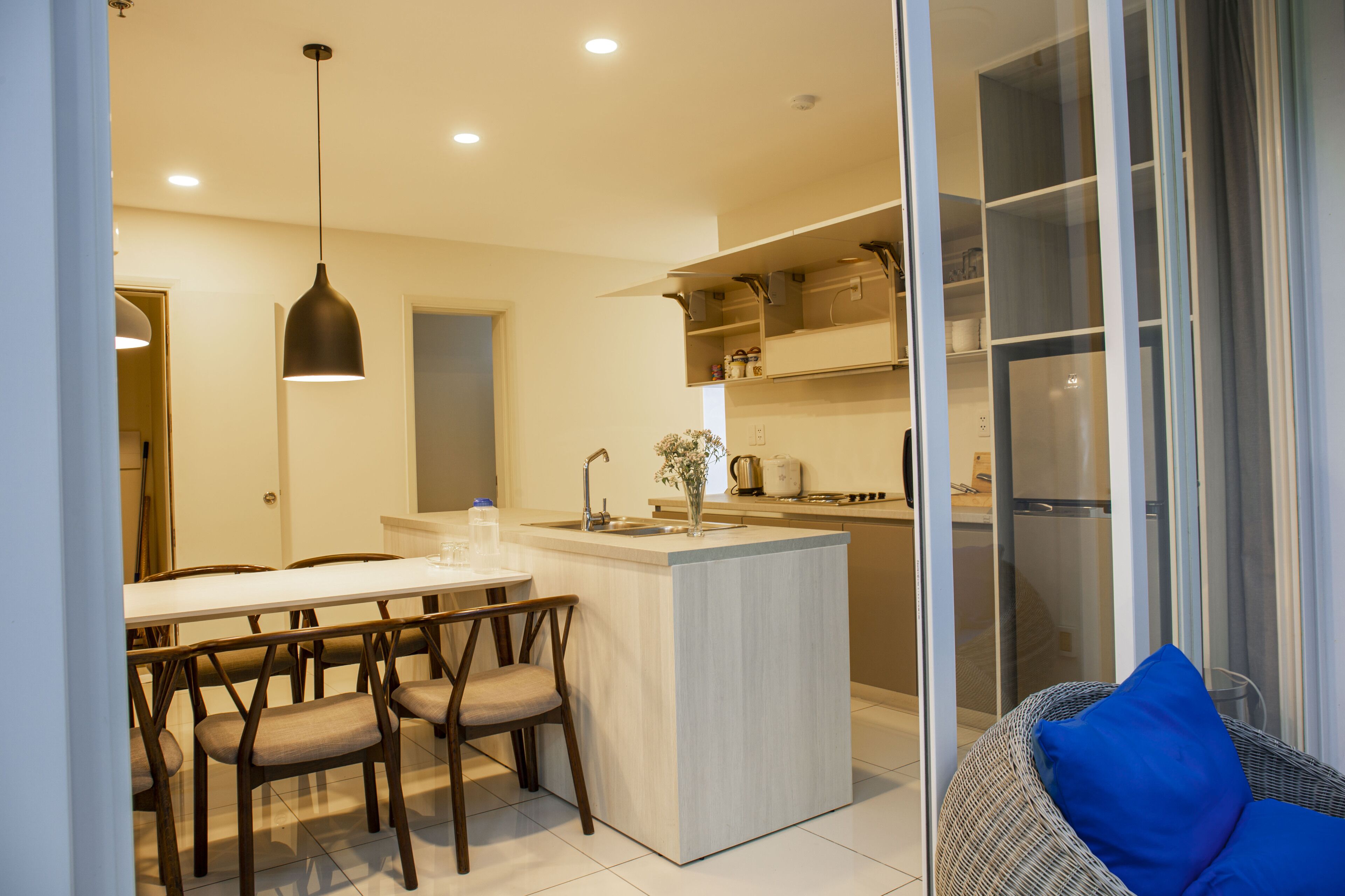 A modern kitchen with a central island, dining area with chairs, and a wicker chair with a blue cushion.