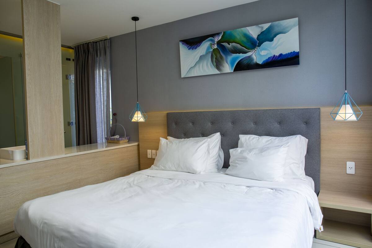 Un lit bien fait, avec du linge de lit blanc, une tête de lit rembourrée de couleur grise et un luminaire suspendu au design moderne.