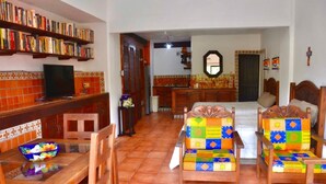 A living room with a TV, bookshelves, a dining table with chairs, a bed with colorful bedding, and a kitchen area in the background. - Beach FrontFully  Equipped Suite # 4 (Centro - Supmza. 001)