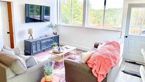 Un salon comprenant un canapé, une table basse, un téléviseur fixé au mur et de grandes fenêtres donnant sur un paysage verdoyant.