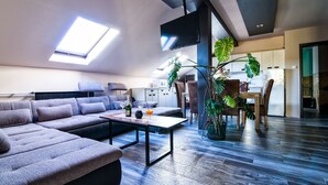 A modern living room with a sectional sofa, a coffee table, and a dining area with chairs and a table. There is a large plant in the center, a ceiling fan, and a TV mounted on the wall. - Thermal Panzió Sárvár (Sarvar)