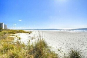 A beach with sand and grass, a clear sky, and buildings in the distance. - Perfect Vacation Getaway! Beach Condo in Quiet Setting, CLOSE TO THE BEACH! (Myrtle Beach)
