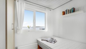 Una habitación pequeña y luminosa con una cama individual, una ventana que da a una masa de agua y una estantería con libros.