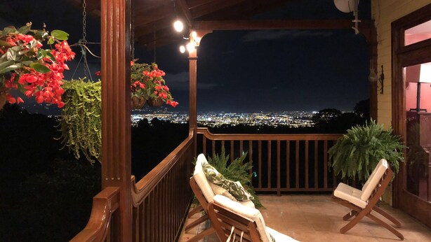 A balcony with two lounge chairs, a hanging plant with red flowers, and a cityscape view at night. - Stunning View of San Jose-private property on 2 acres, Casa Los Cielos & Studio  (Mata de Plátano)