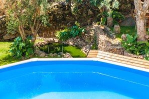 A swimming pool with a wooden deck, surrounded by greenery and a stone wall.