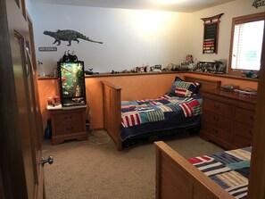 Une chambre à coucher comprenant un lit, une commode, une étagère avec divers objets, un autocollant mural représentant un dinosaure et une fenêtre avec des stores.
