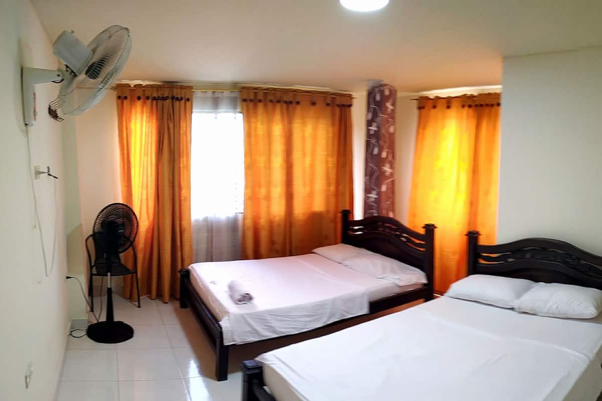 A room with two single beds, a standing fan, and a window with curtains.