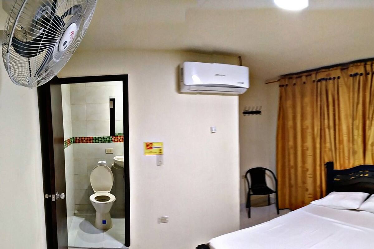 A hotel room with a bed, a fan, an air conditioner, a toilet, and a chair.