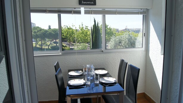 A dining area with a table set for four, a window with a 'bienvenue' sign, and a view of trees and buildings outside.