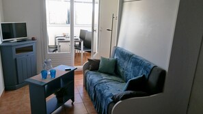 Un pequeño espacio de sala con un sofá, un televisor sobre un mueble, una mesa con bebidas y vistas al exterior.