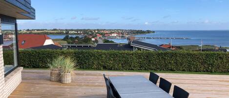 Una terraza de madera con una mesa y sillas, con vistas a un pueblo costero con casas y un puerto deportivo.